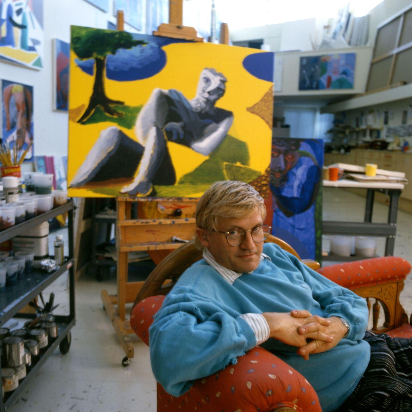 David Hockney seduto nel suo studio con maglione azzurro e dipinti alle spalle.